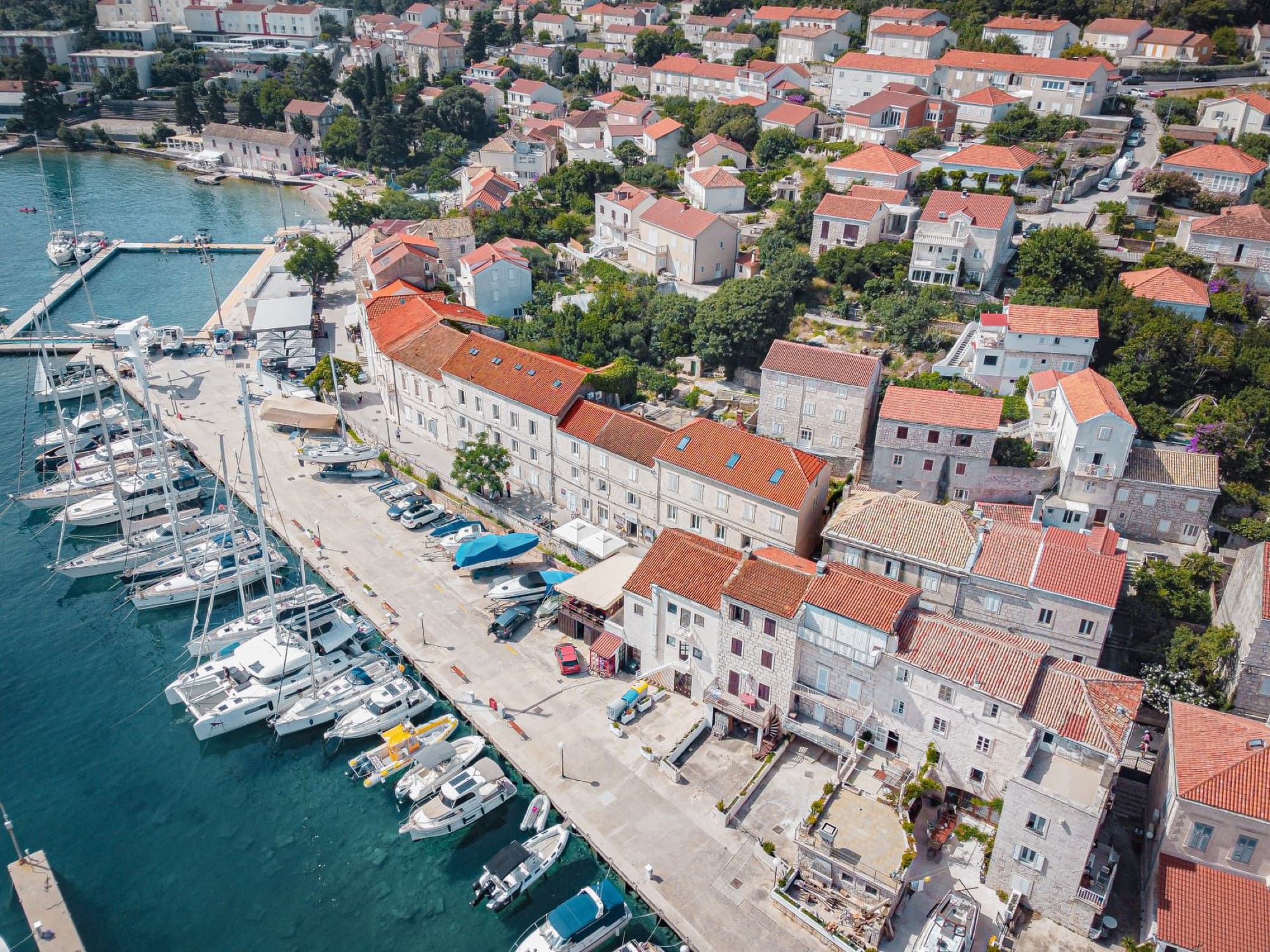 Aerial shot of a marina with yachts, red-roofed buildings in a coastal town.